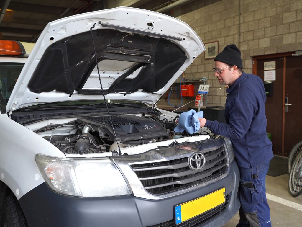 Man gekleed als monteur werkt aan auto met openstaande motorkap.